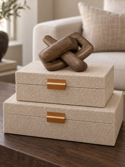 Två beige linneklädda förvaringsboxar med mässingsfärgade beslag, staplade med en brun träknut ovanpå.